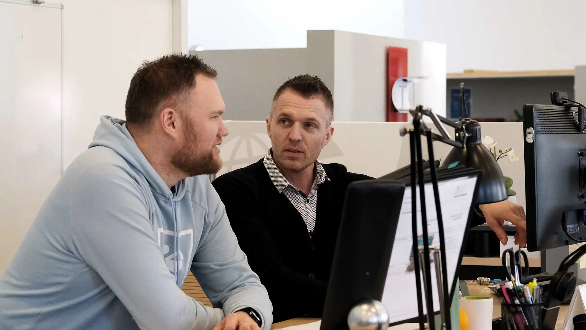 Two men standing at a desk talking and point at a computer screen