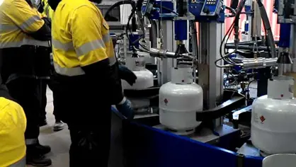 image of 2 workers wearing yellow safety jackets handling a blue filling carousel filling white LPG cylinders
